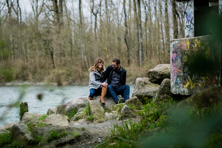 couple holding hands and sitting on rocks in front of the fraser river