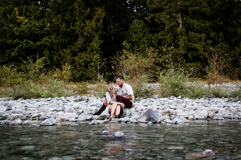engaged couple sitting on the stoney shore of a creek in dewdney bc